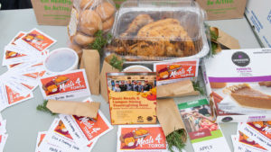 A full cooked turkey, boxed pumpkin pie, rolls, and sprigs of fresh herbs on a table with donation cards 
