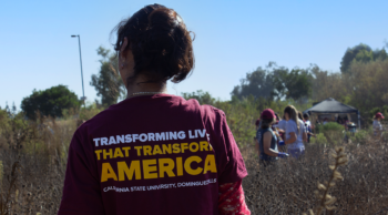 CSUDH student wearing a Tshirt that says "Transforming Lives that Transform America" faces away from the camera stands in a field. Group of people stand in the background.
