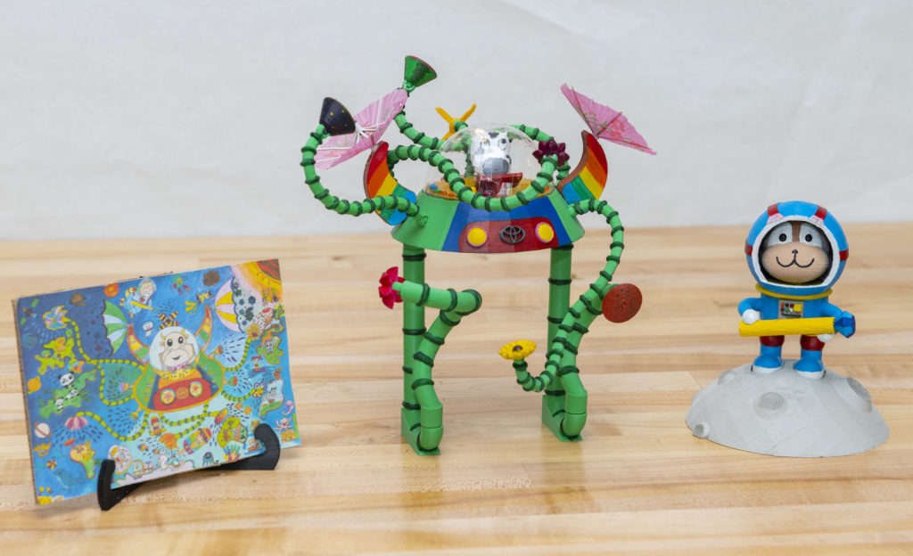 3D printed figures, one of a dog in a blue spacesuit, next to a green vehicle on stilt letgs and tenticle-like arms, driven by a cow. To the left, the child's drawing that inspired the model.