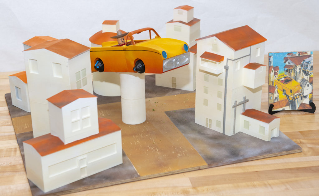 A 3D printed model of of a yellow-orange 3D printed car "flying" through a 3D-printed town, with the drawing that inspired it in a stand to the right of the model.