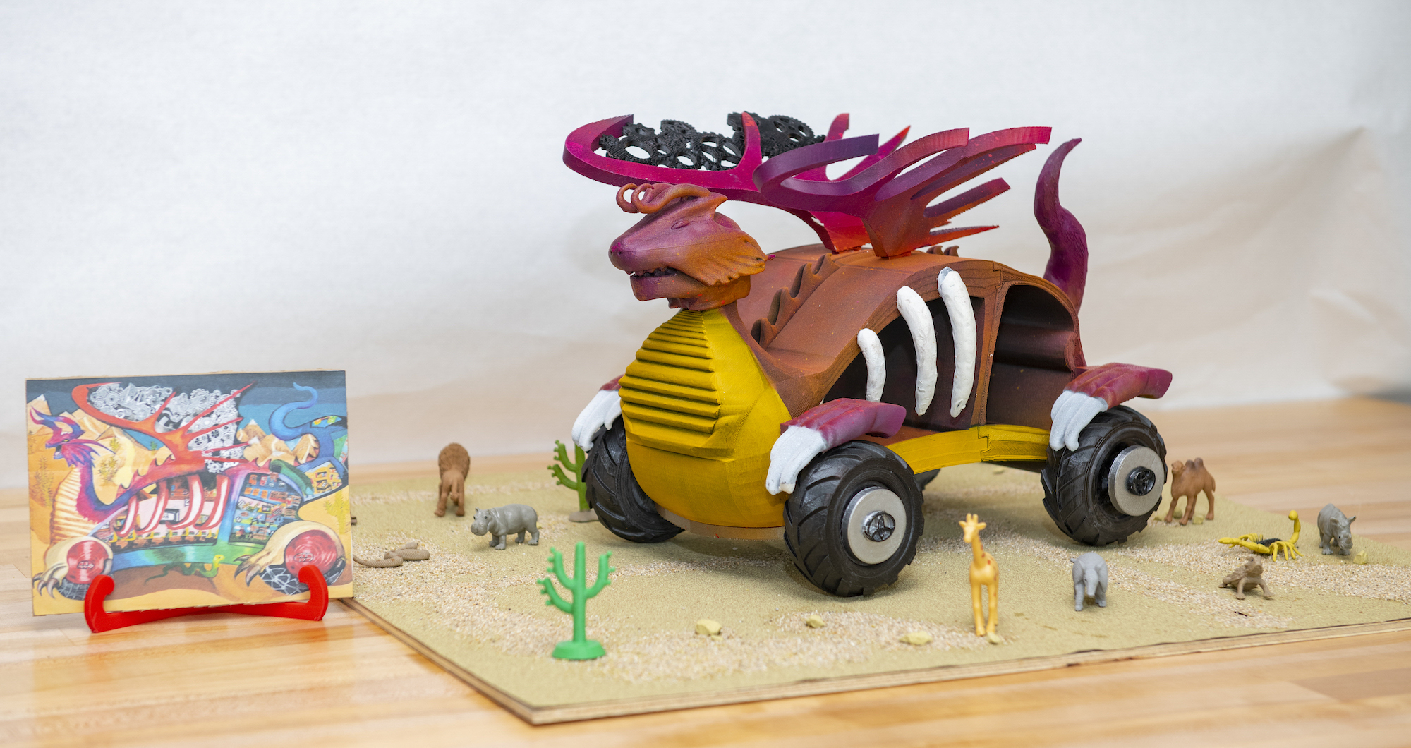 A 3D-printed model of a dragon car on a desert landscape. Next to it is the child's drawing that inspired it.