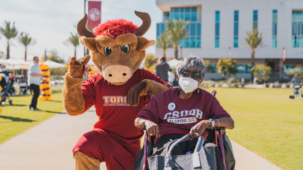 The CSUDH bull mascot kneels down next to a woman in a wheelchair.