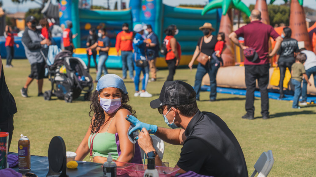 A person smiles under their mask ask a vendor applies a temporary tattoo. A number of people area walking in the background checking out inflatable games and rides.