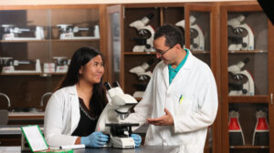 Two people wearing white lab coats in a laboratory, with a microscope