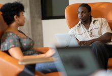 Two individuals are seated in orange armchairs, engaged in conversation while one uses a laptop.