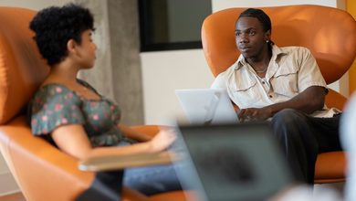 Two individuals are seated in orange armchairs, engaged in conversation while one uses a laptop.